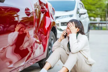 A frustrated woman sitting on the ground beside her dented car after a crash, reflecting the question many drivers ask: do I need a lawyer for a minor car accident?