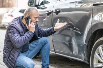 Man standing beside his wrecked car and making a phone call, wondering is uninsured motorist coverage required in California.