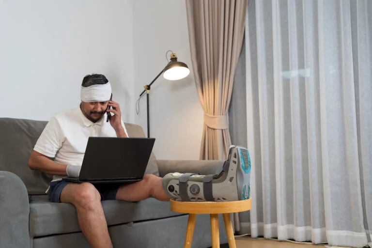 An injured man wearing a walking boot and head bandage talks on the phone with his lawyer, representing clients asking why is my car accident settlement taking so long.