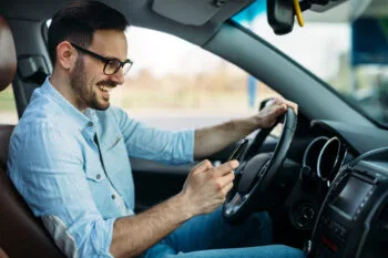 A man laughs while looking down at his smartphone behind the wheel, showing risky behavior restricted under the California No-Touch phone law.