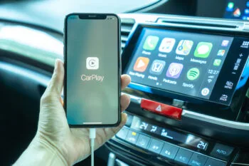 An iPhone displays the Apple CarPlay app next to a car dashboard screen, connecting to the topic can you watch movies on Apple CarPlay while driving in California.