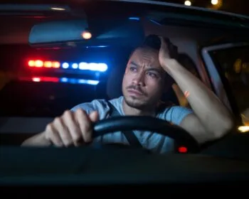 A man sits in his car looking frustrated as police lights flash behind him, depicting driving with a suspended license in California.