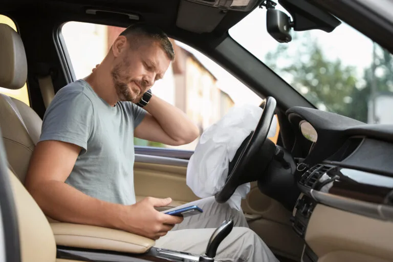 A man sits in his car holding his shoulder in pain after a crash with airbags deployed, representing an insurance lapsed car accident.