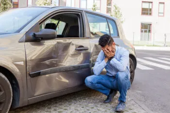 A person bends down inspecting damage to their vehicle, representing frustration after someone hit my parked car and left without leaving a note.